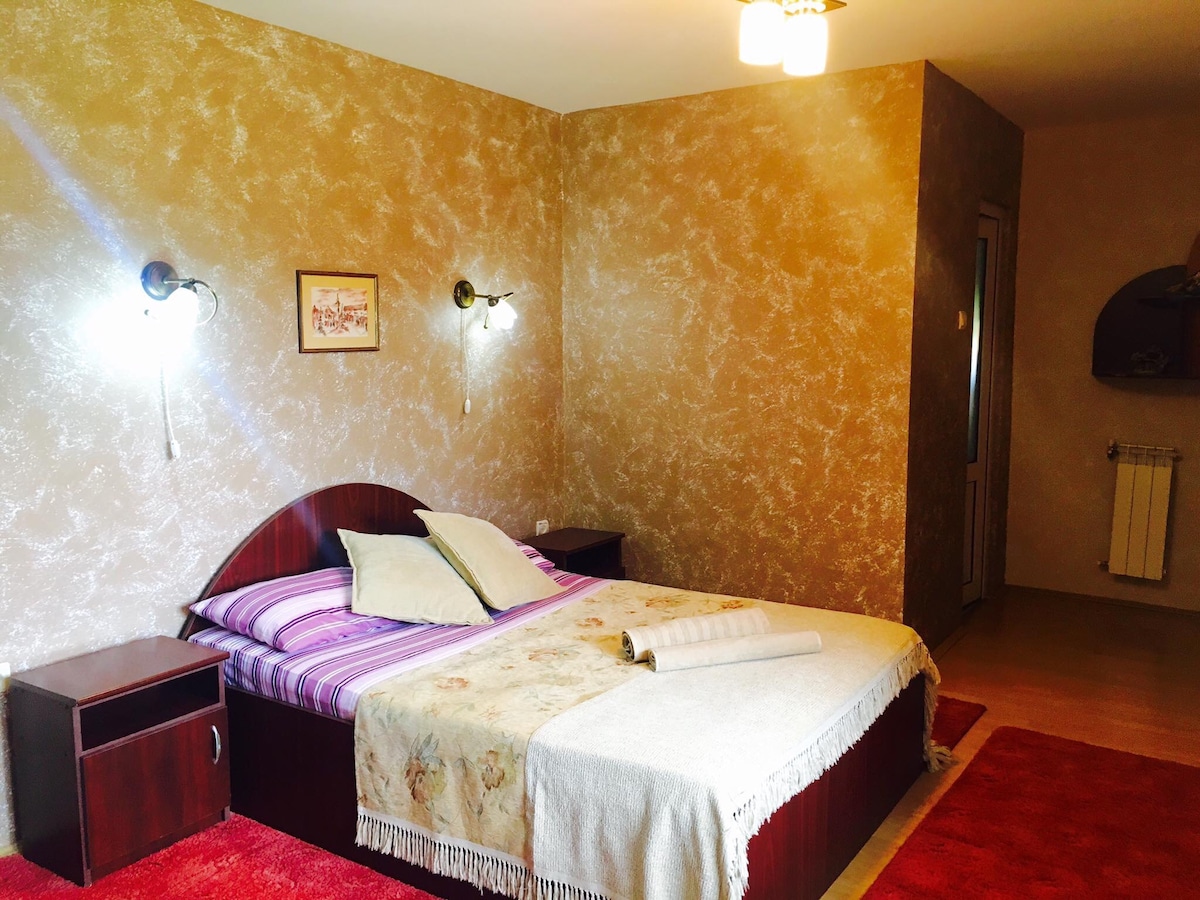 A spacious sleeping area features a large bed with a decorative blanket and pillows. Soft lighting is provided by wall-mounted sconces on either side. A small nightstand sits adjacent to the bed, and warm tones of the walls create a calming environment.