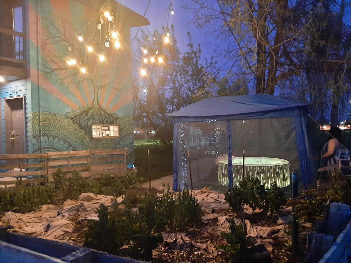 An outdoor area is illuminated by string lights, casting a warm glow over a cozy seating arrangement under a canopy. An edible garden with neatly arranged plants is visible in the foreground, accompanied by a vibrant mural on the adjacent building.