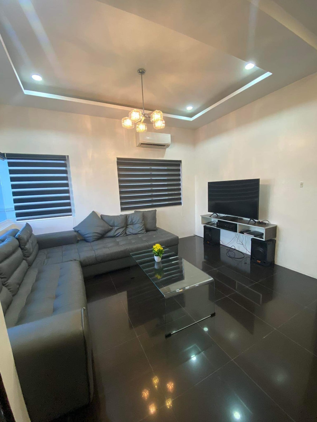 A modern living area features a sectional sofa and a glass coffee table. Dark blinds cover the windows, allowing for adjustable light. A television is mounted on the wall opposite the seating, complemented by a console below. A small vase with a flower adds a touch of color.