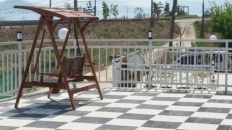 An outdoor terrace features a checkered tile floor and a wooden swing, offering a relaxing seating option. The area is well-lit with lamp posts, and the surrounding landscape includes green fields and distant mountains under a clear sky.