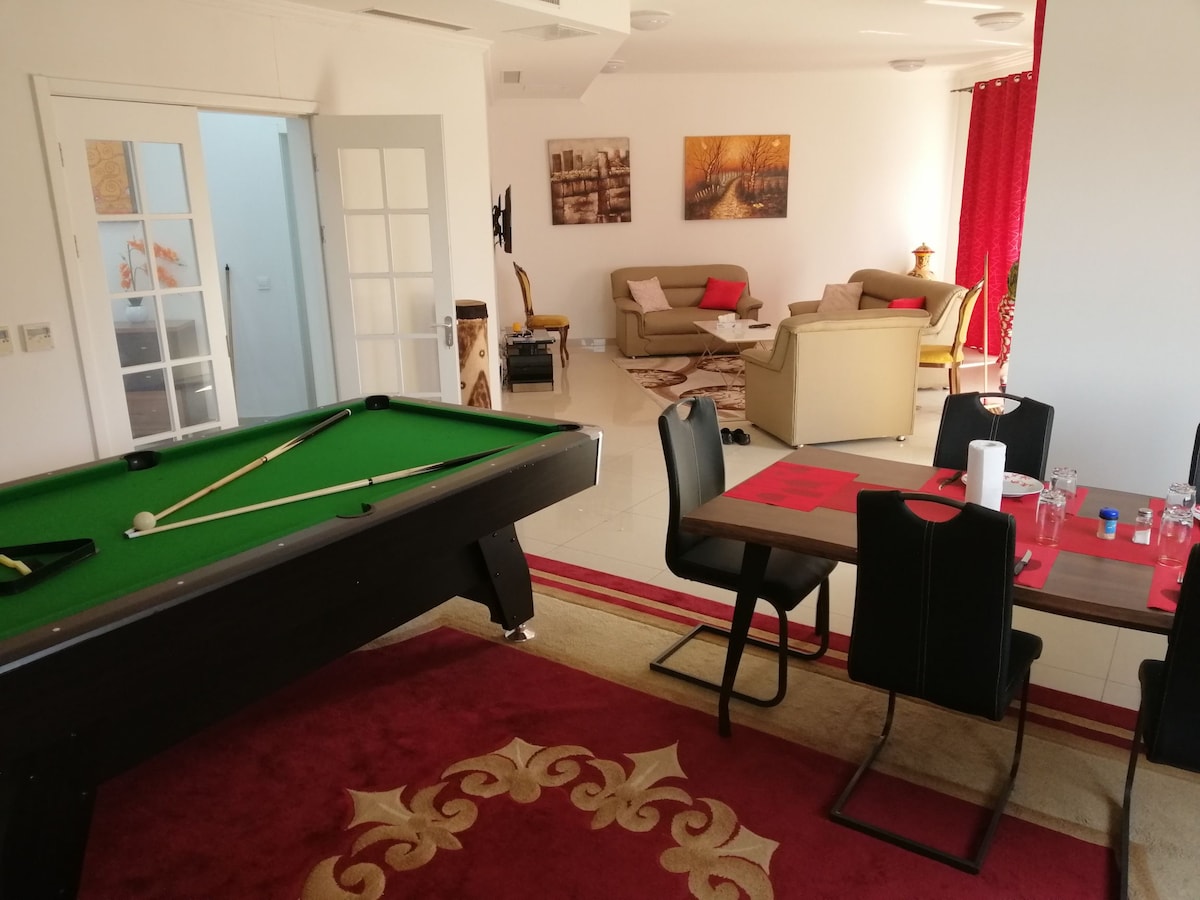 An open living area features a green pool table alongside a dining table set for six. A cozy seating arrangement is visible with neutral-toned sofas, and red drapes add a touch of color. Natural light flows through large windows, illuminating the space.