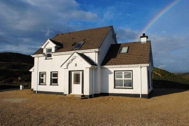 The holiday home is a two-story structure with a simple white facade, accented by a grey roof and bordered windows. A rainbow arcs above, contrasting with the blue sky. A gravel area surrounds the house, providing ample space.