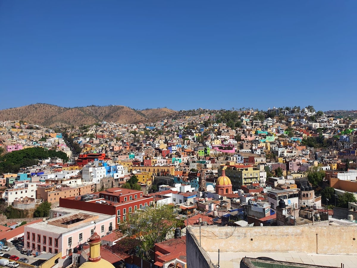 A panoramic view of Guanajuato reveals a vibrant tapestry of colorful buildings nestled among rolling hills. The scene highlights the diverse architecture, with rooftops in various shades, and a distinctive dome stands out amidst the cityscape under a clear blue sky.