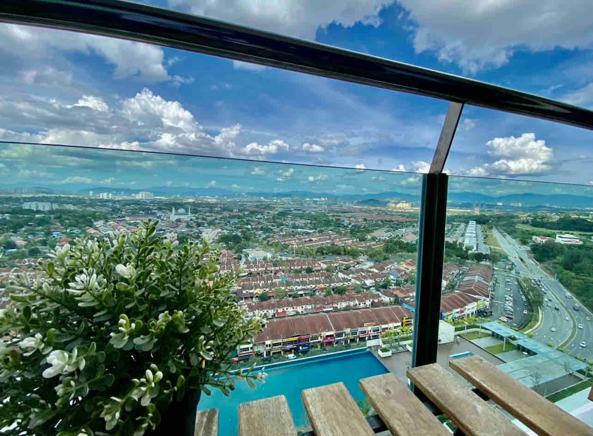 A spacious balcony is presented with glass railings, offering a panoramic view of the surrounding town and distant hills. A small potted plant adds a natural touch, while the sky is adorned with fluffy clouds, creating an airy and open atmosphere.