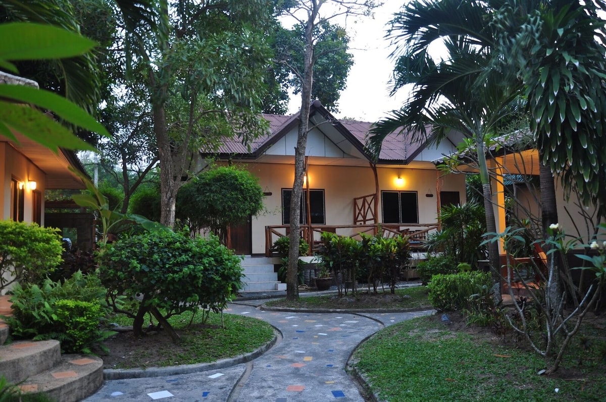 Traditional bungalows are situated within a lush tropical garden, surrounded by pathways and greenery. The serene environment features well-maintained plants, with soft lighting illuminating the bungalows in the early evening. Varied foliage creates a tranquil and inviting atmosphere.