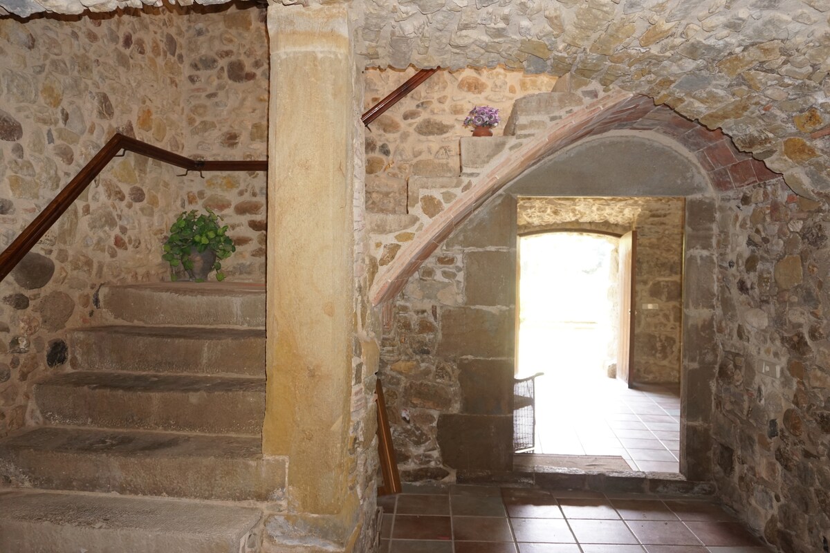 An entryway is framed by rustic stone walls, showcasing a blend of historical charm and natural light. A staircase with a wooden handrail leads upward, while an open doorway at the far end invites views of the garden outside. A small potted plant adds a touch of greenery.