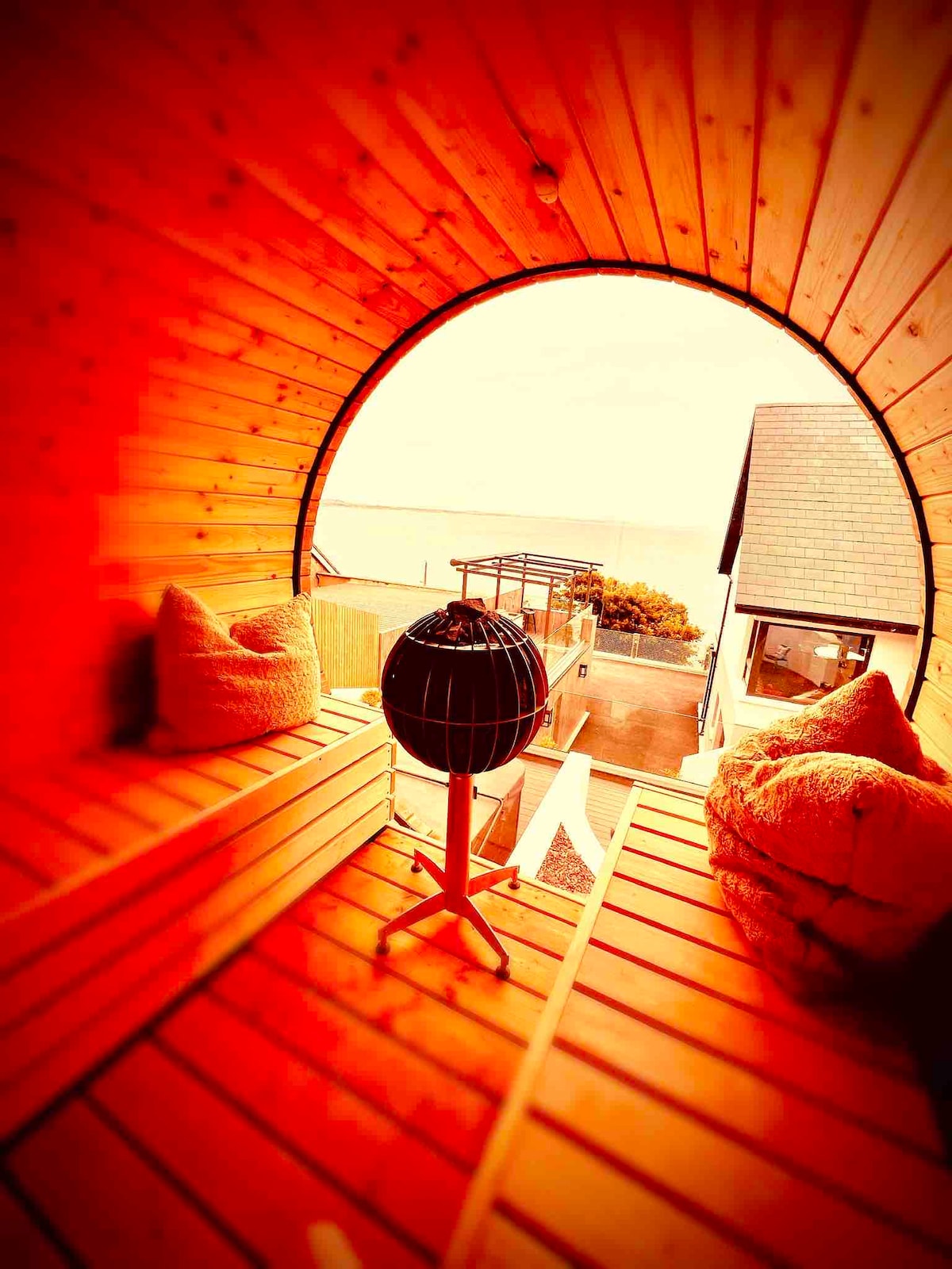 A cozy seating area is enclosed by warm wooden walls, featuring plush cushions arranged comfortably. A circular window offers stunning views of the sea, while a unique spherical light fixture adds character to the space. The ambiance is inviting and stylish.