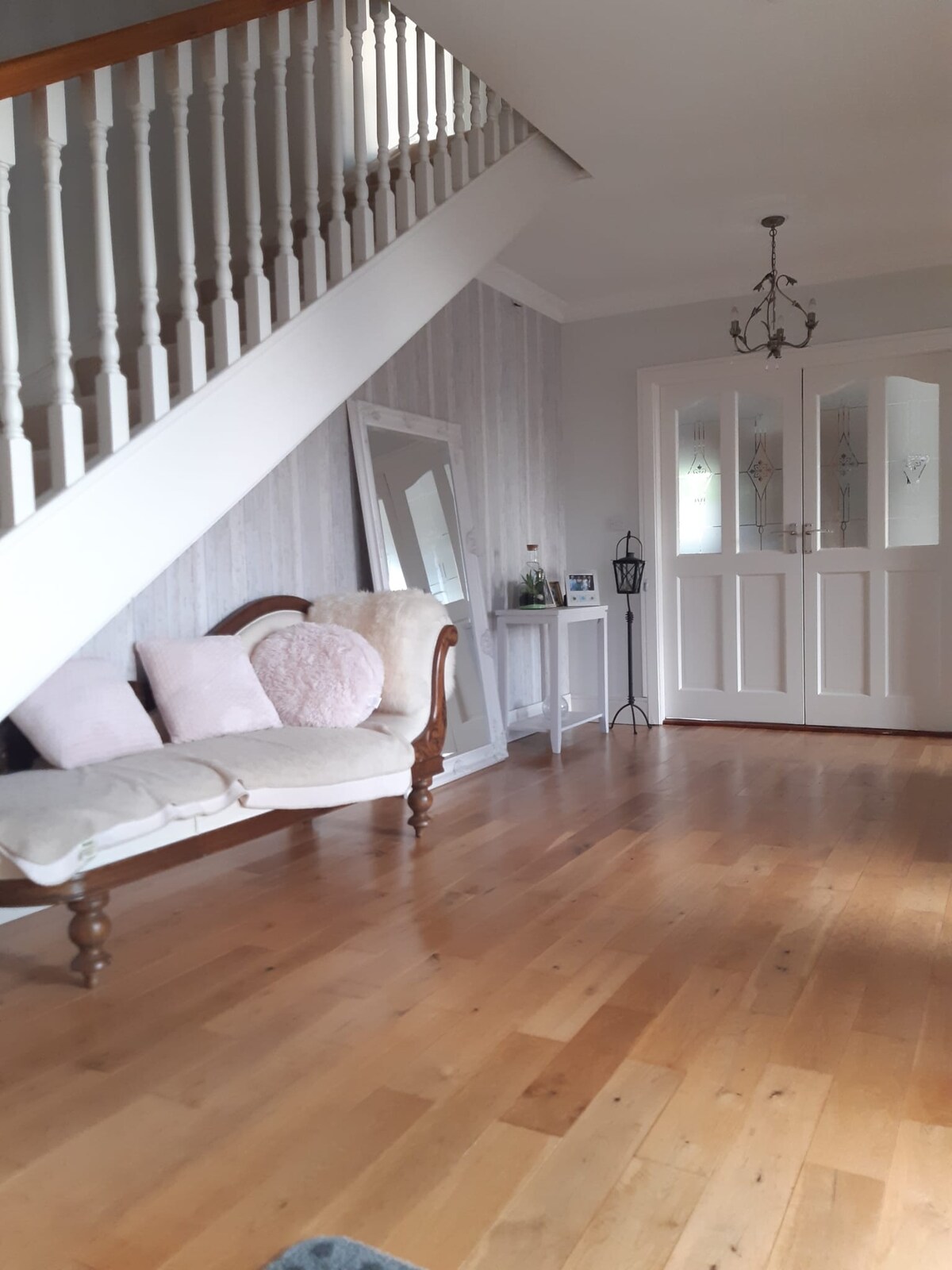 A welcoming entrance area features a light wooden floor beneath a staircase. An elegant vintage-style sofa adorned with soft pillows is placed against the wall, beside a decorative mirror. Double doors with glass panels lead to another room, enhancing the space's openness.