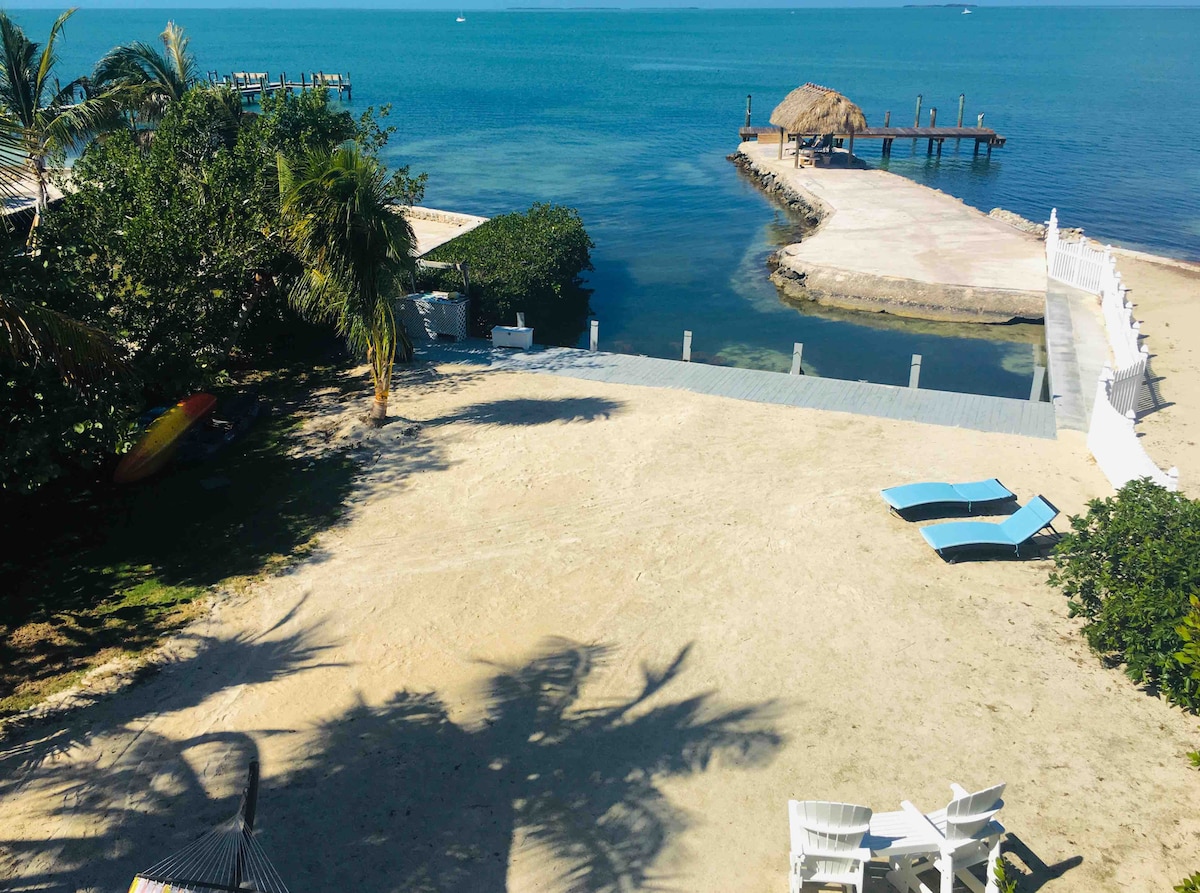 A sandy beach area extends towards calm blue waters, featuring lounge chairs positioned for relaxation. A private dock is visible, alongside the jetty, leading into the water. Lush greenery surrounds the space, enhancing the tranquil coastal setting.