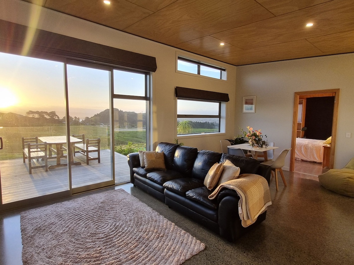 A spacious living area features a comfortable black leather sofa accompanied by a textured area rug. Large glass doors allow natural light to fill the space and offer a view of the outdoor dining area. Light wooden panels create an inviting atmosphere.