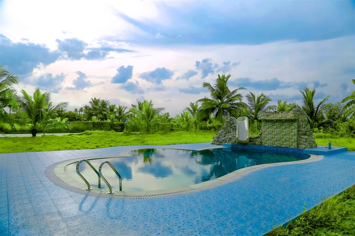 A serene swimming pool is surrounded by a lush green lawn, with palm trees framing the view. The pool features a smooth blue tile surface and two steps leading into the water. The backdrop displays a cloudy sky, enhancing the tranquil atmosphere.