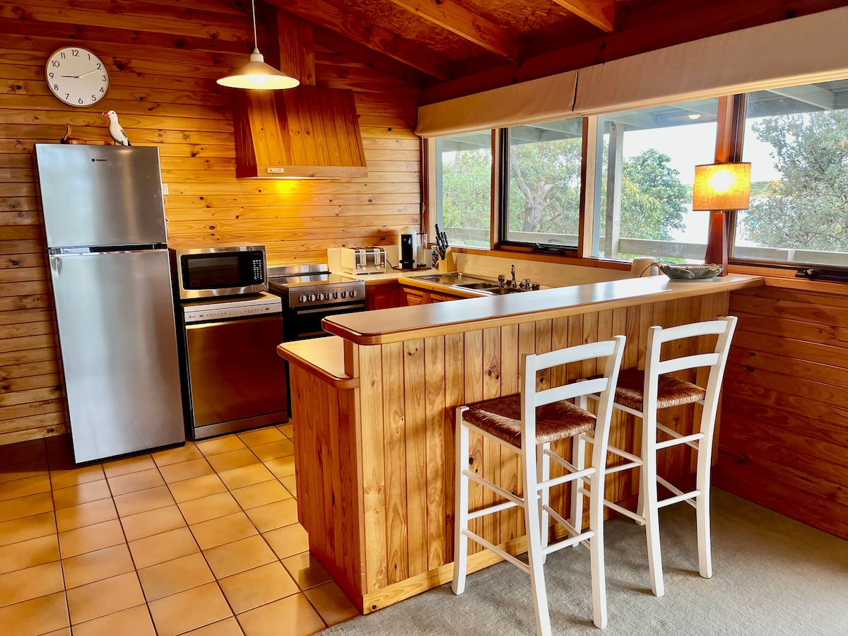 The kitchen features warm wooden paneling with ample natural light from large windows. Modern appliances include a refrigerator, microwave, and stove. Two white stools are positioned at the wooden breakfast bar, which faces an open cooking area, creating an inviting space for meal preparation.