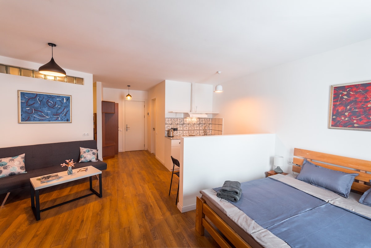 A cozy living space is comprised of a double bed with soft bedding, a sofa with a small coffee table, and dining area featuring a kitchenette. Artwork adorns the walls, and warm wooden flooring complements the inviting atmosphere.
