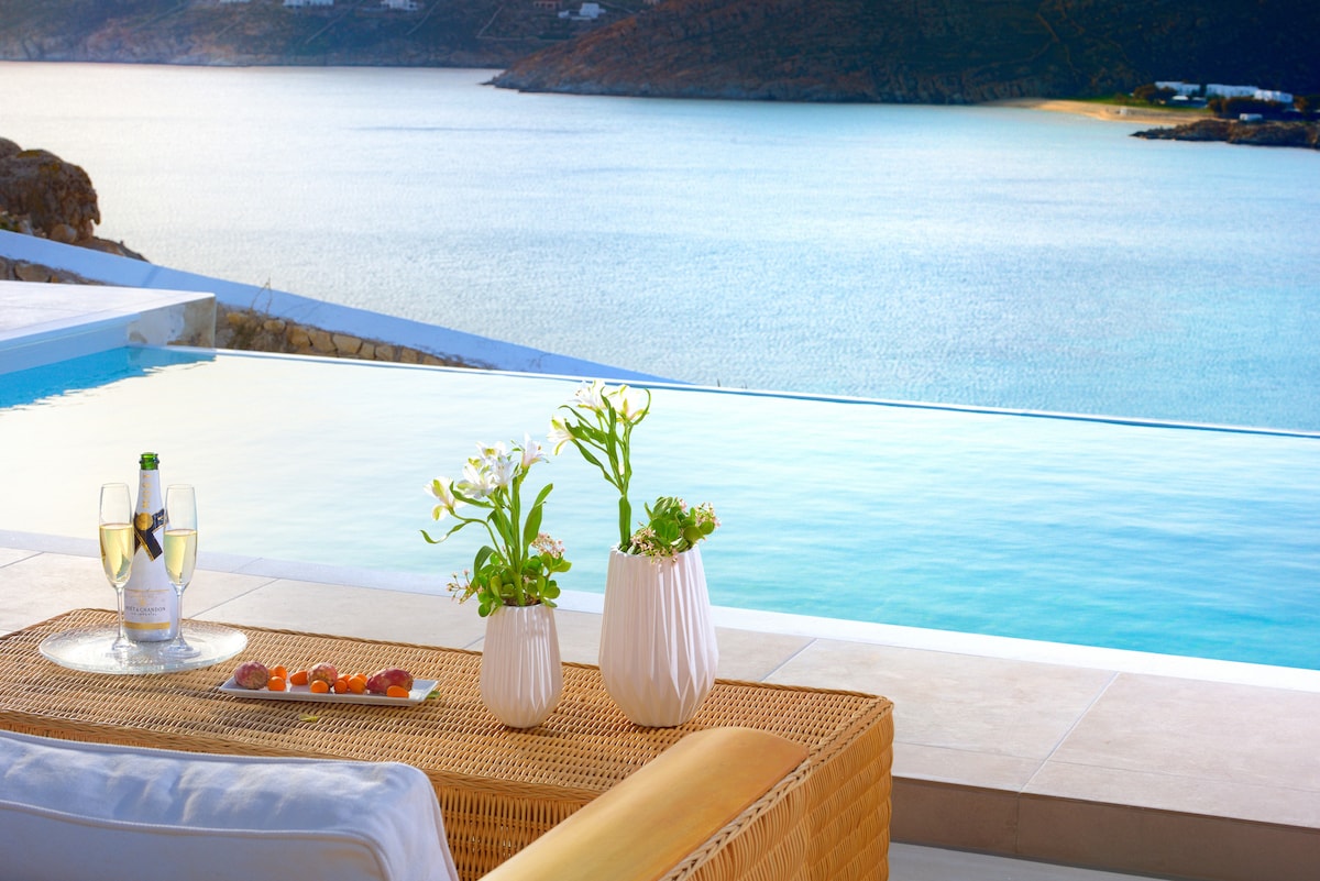 A serene outdoor setting features a modern seating area overlooking a tranquil infinity pool. Two vases with fresh flowers are placed on a decorative table, alongside a plate of fruit and two glasses filled with sparkling beverage. The sparkling sea and distant coastline are visible in the background.