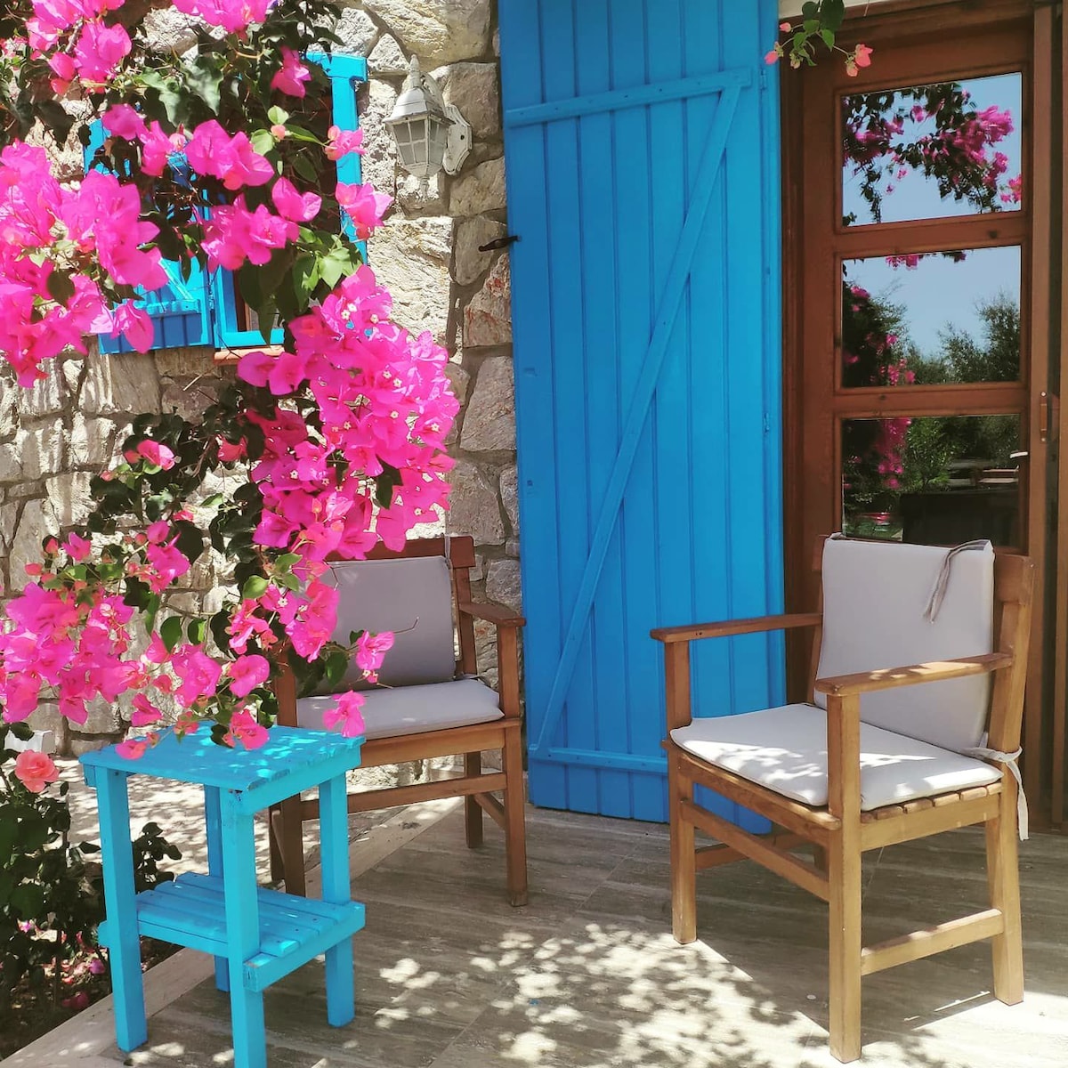 A charming outdoor seating area is highlighted by two wooden chairs with cushions, accompanied by a small blue table. Vibrant pink bougainvillea cascades over the stone wall, while a bright blue door adds a pop of color to the serene setting.