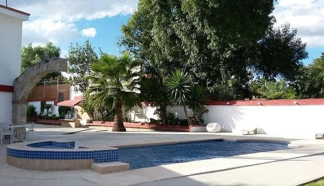 A spacious outdoor area features a clear swimming pool surrounded by tropical plants. The pool is complemented by a shaded seating area and a stone archway. Sunlight creates a bright atmosphere, highlighting the well-maintained landscaping and inviting space for relaxation.