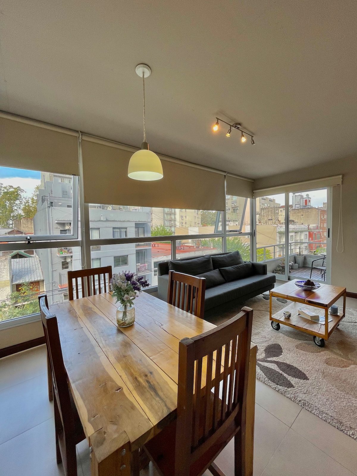 A dining table with wooden chairs is positioned near large windows allowing natural light to fill the space. A comfortable sofa is visible in the background, complemented by a soft area rug. Decorative elements include a vase of flowers on the table.