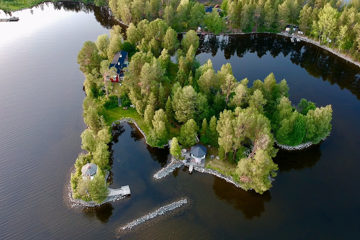 An aerial view captures the serene private island of Linnéaholmen surrounded by calm waters. Lush greenery and trees encircle the property, while pathways lead to different structures, including a sauna house. The peaceful setting is accentuated by the reflective surface of Storsjön.