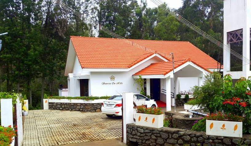 The exterior of a luxury guest house is shown, featuring a sloped terracotta roof and white walls. A paved driveway leads to the entrance, surrounded by well-maintained greenery and decorative plants. Ample parking space is visible in front, accommodating vehicles.