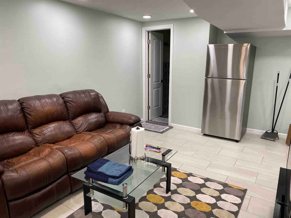 A living room is featured with a brown leather sectional sofa positioned next to a glass coffee table. A silver refrigerator is visible, along with a doorway leading to the bathroom. Light-colored walls and tiled flooring enhance the room's spacious feel.