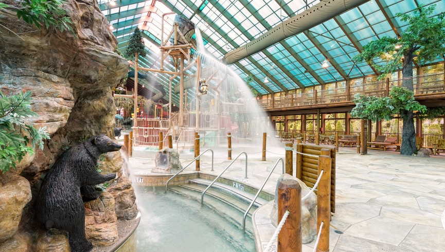 An indoor water park features a large, rocky structure with a bear sculpture. Water cascades from heights, creating an inviting atmosphere. Surrounding the water area, wooden posts and natural foliage can be seen, while nearby benches provide seating for relaxation.