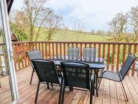 An outdoor dining area is presented on a wooden veranda, featuring a black glass-top table surrounded by six black mesh chairs. Trees and green fields are visible in the background, providing a natural setting.