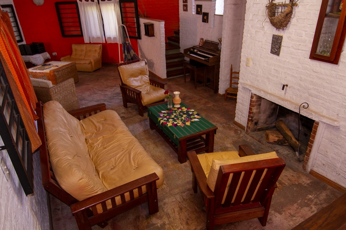 A cozy living area features a brown leather sofa and matching chairs arranged around a green table, adorned with colorful decor. Natural light enters through windows, illuminating the rustic brick walls and wooden accents. A staircase is visible in the background, leading to an upper level.