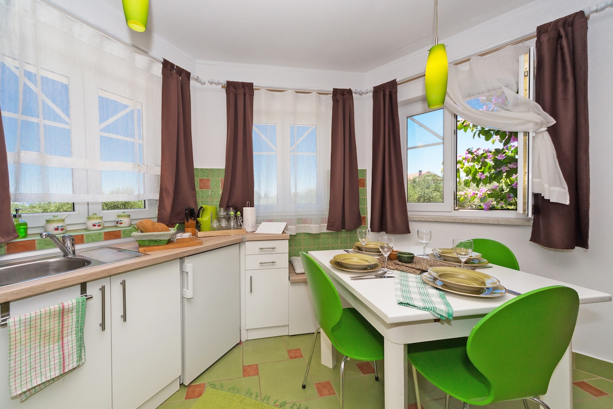 The kitchen area features a clean and functional layout, with white cabinets and a stainless-steel sink. A dining table with vibrant green chairs is set for meals, illuminated by pendant lights. Large windows provide natural light and views of the surrounding landscape, framed by light curtains.