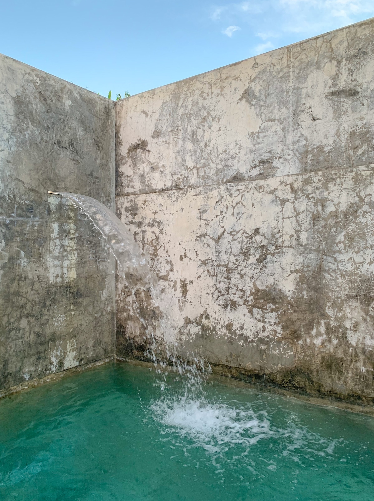 A water feature is observed cascading gently from a wall into a serene pool, creating small ripples on the surface. The raw cement walls exhibit unique textures, contributing to the natural aesthetic of the space. The water's clear turquoise color enhances the tranquil atmosphere.