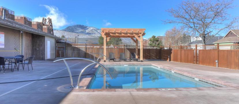 A clear view of a pool area is presented, featuring a sunlit swimming pool with a gentle water feature. Surrounding the pool are multiple lounge chairs positioned under a wooden pergola. A fence and trees frame the space, with mountains visible in the background.