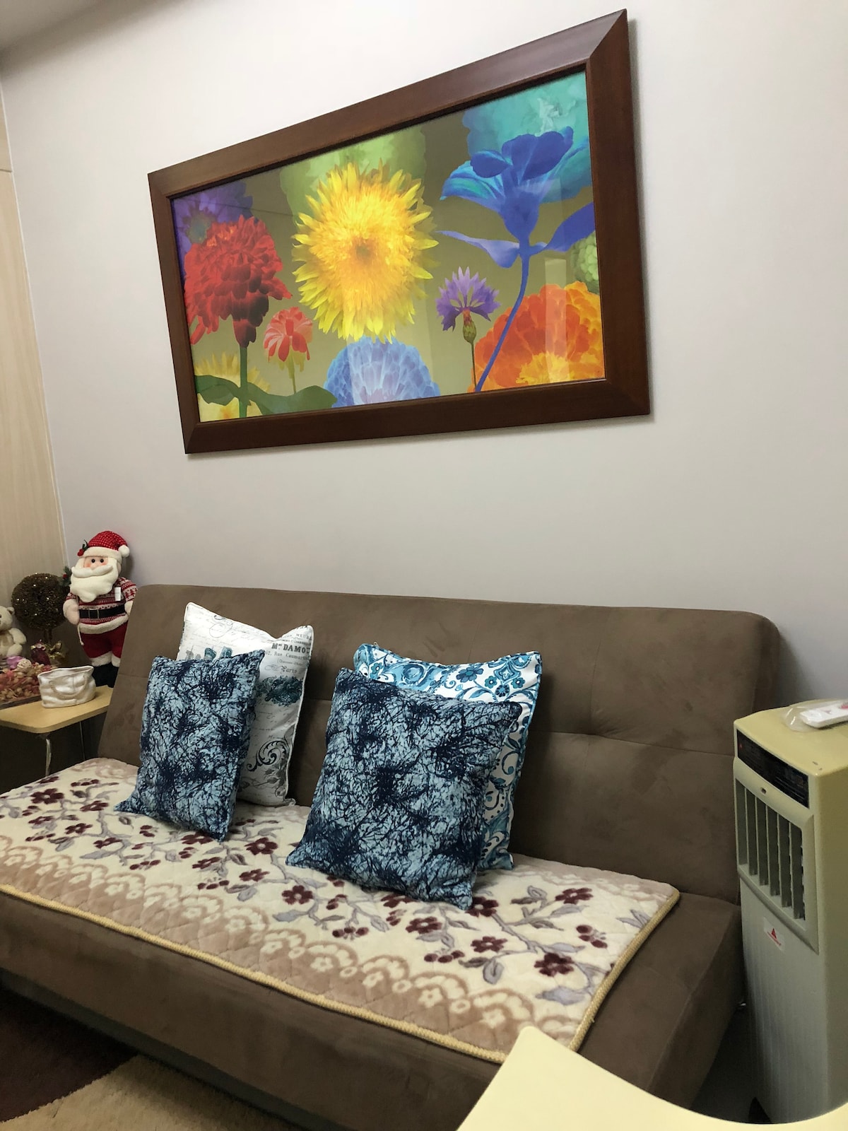 A comfortable sofa bed is placed against a wall, adorned with two decorative pillows featuring intricate blue patterns. Above the sofa, a large framed artwork displays a colorful floral theme. A small side table supports a few decorative items, including a seasonal figure.