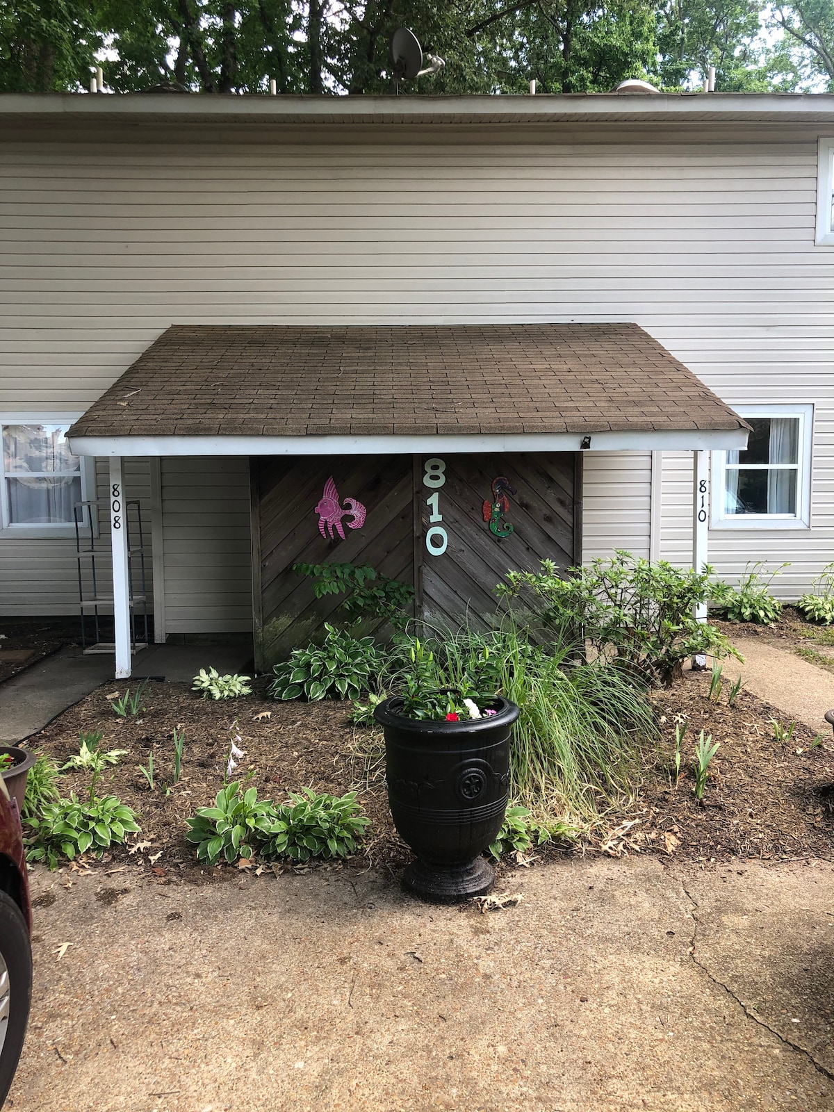 The home’s entrance is marked by a sloped roof and wooden detailing, featuring the number 810 on the door. Lush greenery and flower beds surround the entrance, providing a welcoming touch. A black planter sits prominently in front, contributing to the home's inviting appearance.