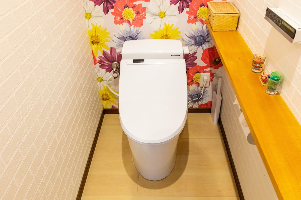A modern toilet is showcased against a vibrant floral wallpaper featuring a colorful design. A wooden shelf on the side holds decorative items. The floor is finished with light-colored wood, contributing to a clean and bright atmosphere.