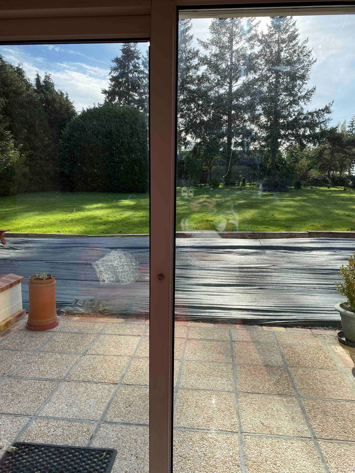A view of the outdoor patio from inside the apartment is displayed, featuring a well-maintained garden with lush green grass and tall trees in the background. Sunlight casts gentle reflections on the glass doors, enhancing the serene atmosphere.
