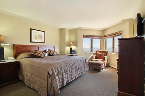 A well-appointed bedroom is equipped with a spacious bed adorned with neutral-toned linens. A comfortable armchair is positioned by the window, which offers views of the outside. Soft lighting is provided by bedside lamps, creating a relaxed atmosphere.