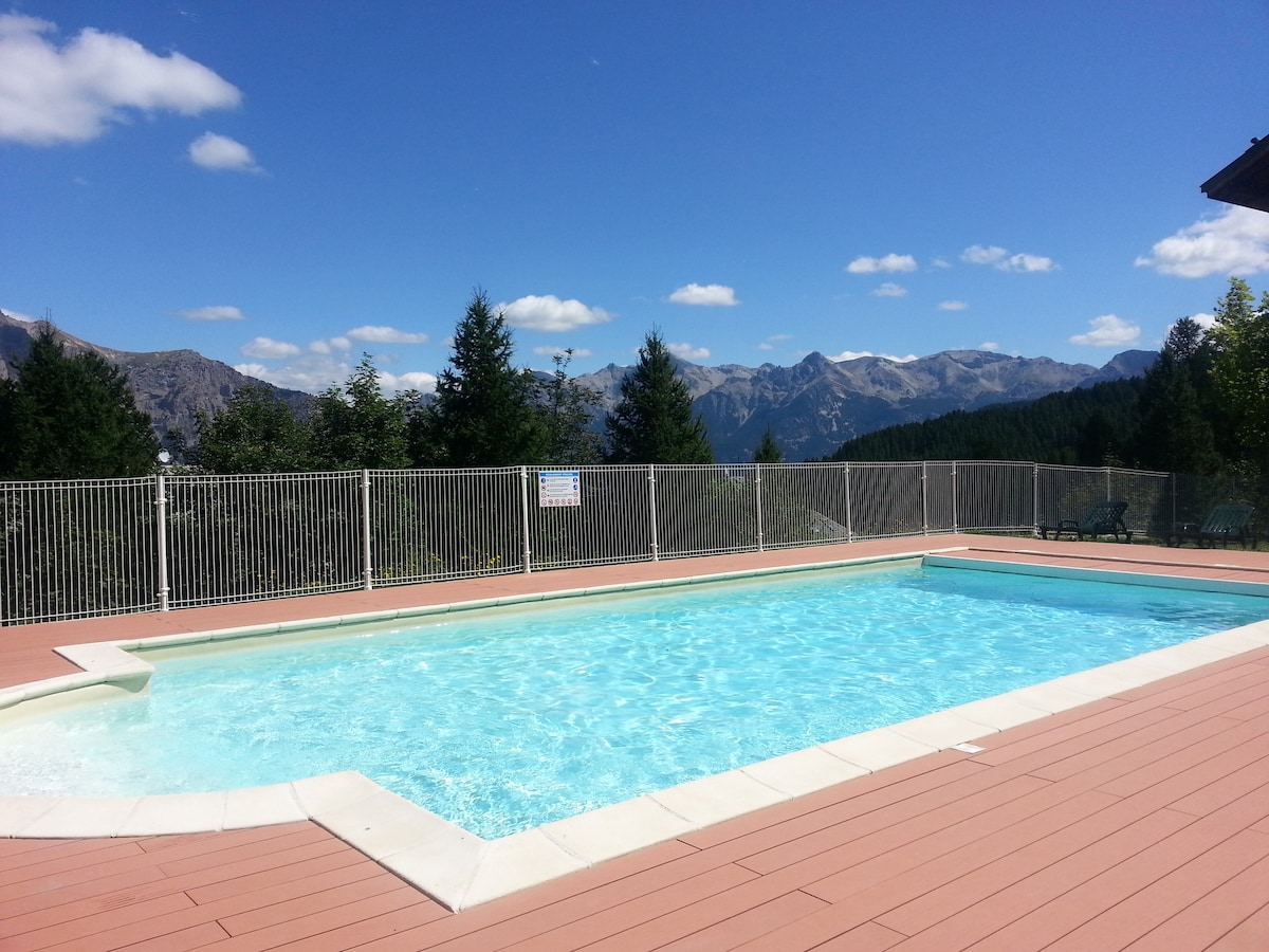 A private outdoor pool is surrounded by a secure fence, providing a clear view of the distant mountains. The pool features bright blue water, inviting for a refreshing swim on warm days. Sunlight illuminates the surrounding deck area, which is complemented by greenery in the background.