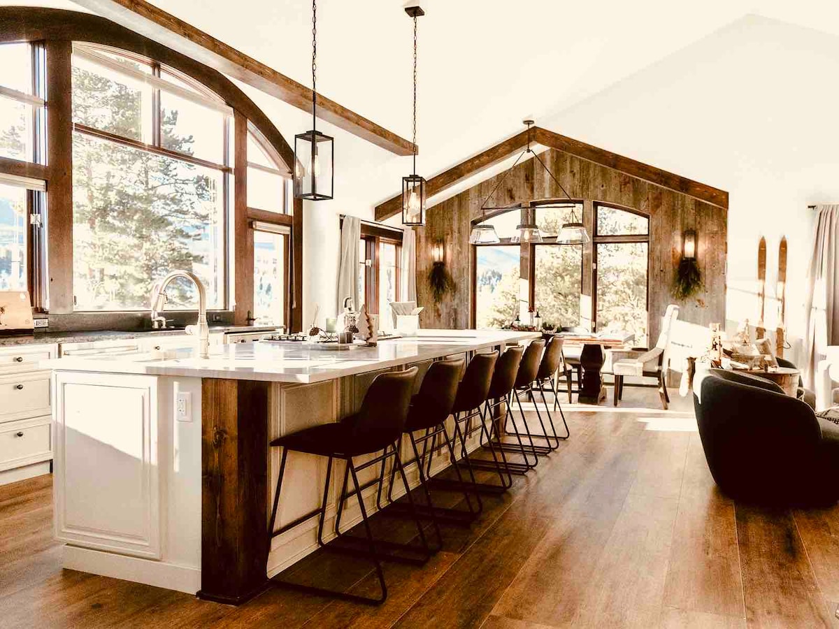 A spacious kitchen is depicted, featuring a large island with seating for several guests. Abundant natural light streams through large windows, illuminating the wood accents and modern appliances. The open layout connects to a cozy living area, all set against a backdrop of trees and mountain views.