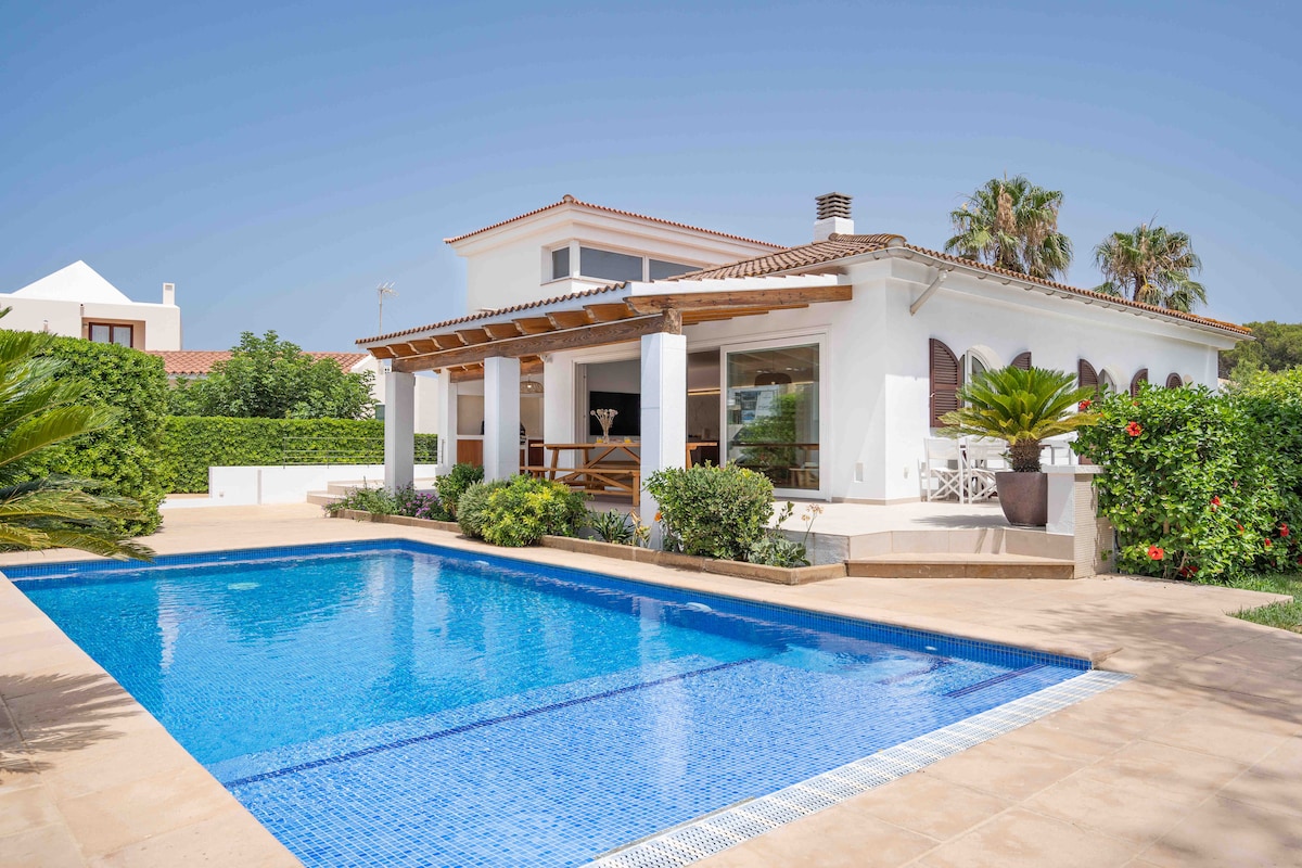 A contemporary villa showcases a spacious outdoor area, featuring a rectangular pool surrounded by a sun terrace and lush greenery. The structure features large windows and a covered porch, creating an inviting space for relaxation. Tropical plants add a vibrant touch to the garden.