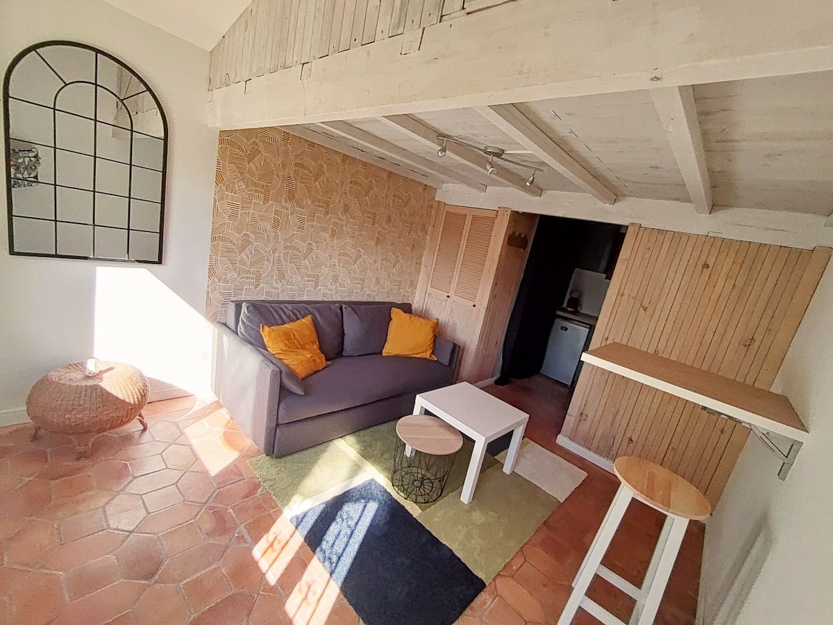 A cozy living area features a gray sofa with yellow cushions, positioned against a textured wall. A small white table sits at the center, accompanied by a round woven stool. Natural light fills the space, highlighting the terracotta floor and wooden beam ceiling.