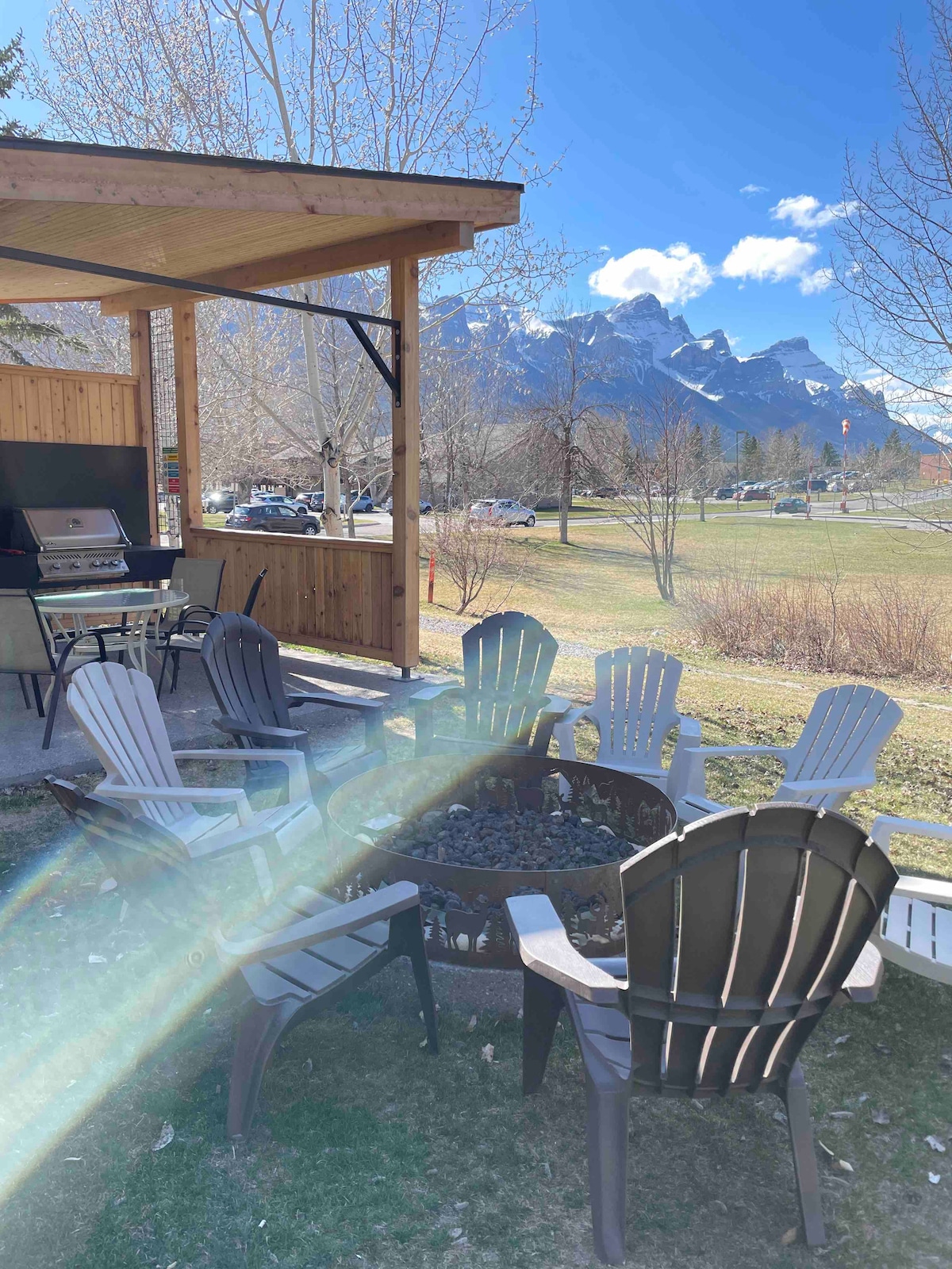 An outdoor seating area features multiple Adirondack chairs arranged around a fire pit, with a landscaped lawn and mountain views in the background. A covered patio area includes a grill, providing a space for outdoor cooking and dining.