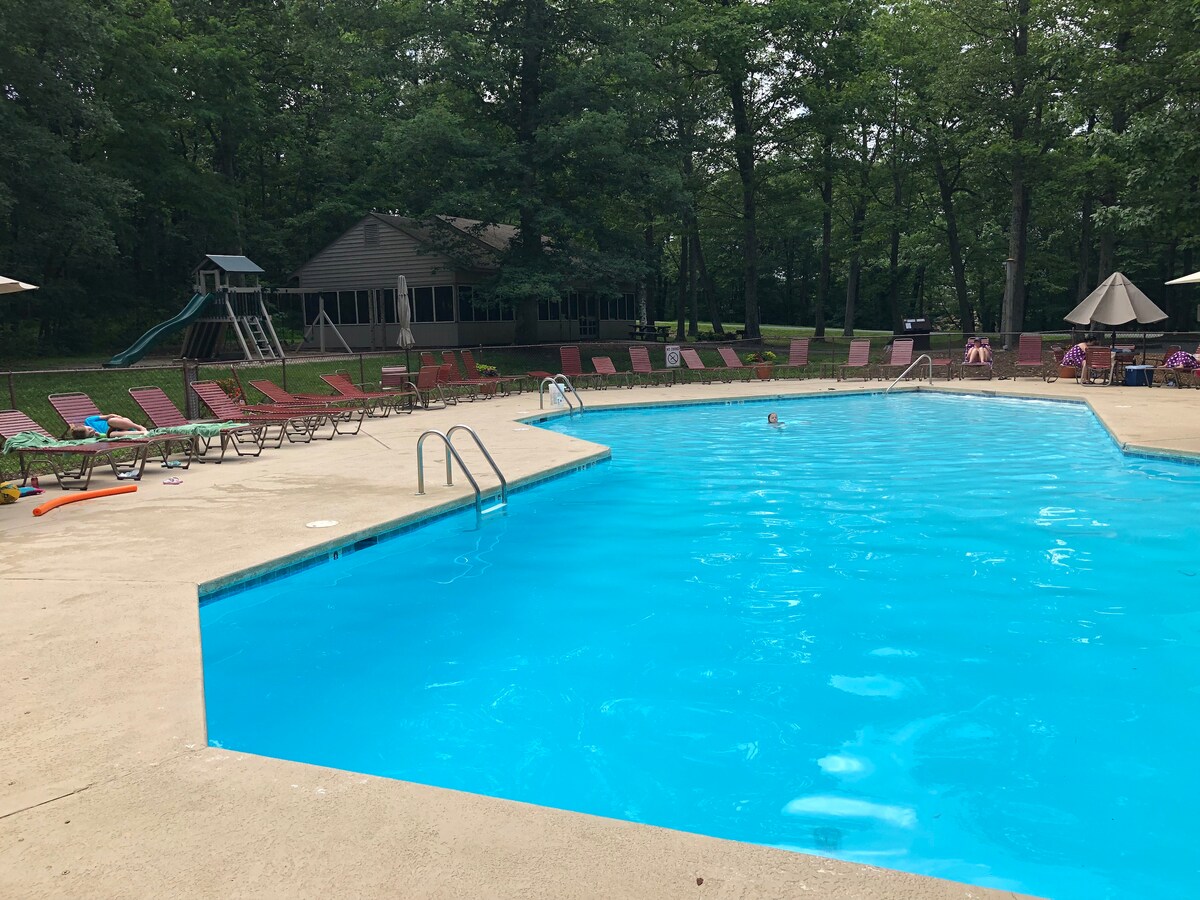 An outdoor pool is surrounded by a spacious deck with multiple lounge chairs. A shaded area with umbrellas provides comfort, while trees line the background, offering a natural setting. A slide structure is visible nearby, enhancing the recreational appeal of the area.