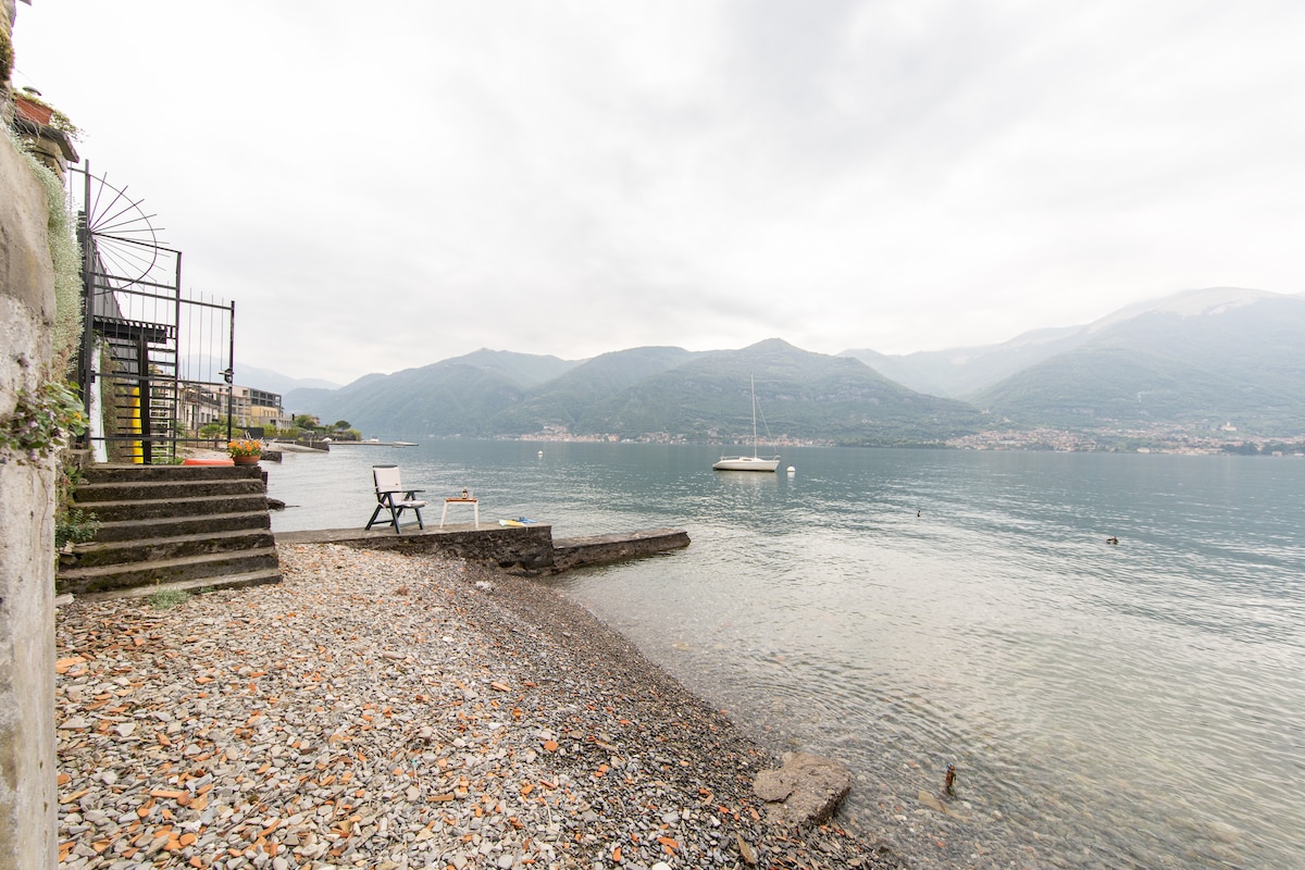 A serene lakeside view features a rocky shoreline leading to calm waters, with mountains in the background under a soft, overcast sky. A wooden chair sits on the shore, and steps can be seen leading to a small terrace overlooking the lake.