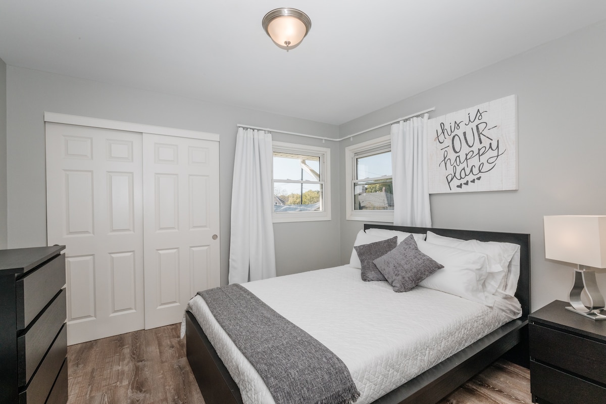 A well-lit bedroom features a spacious bed with neutral bedding and decorative pillows. Two windows allow natural light to enter, and sheer curtains frame the views. A bedside lamp provides additional illumination, while a black dresser and closet doors complete the room's layout.