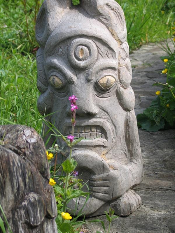 A carved wooden sculpture is positioned along a stone path, surrounded by vibrant green grass and yellow flowers. The natural textures of the wood and the organic setting create a harmonious element within the outdoor space.