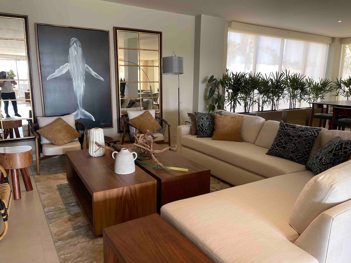 A spacious living area is presented with a neutral color palette, featuring a large artwork of a whale. Comfortable seating arrangements include a light-colored sofa and chairs with accent pillows. Natural light filters through large windows, enhancing the ambiance of the modern decor.