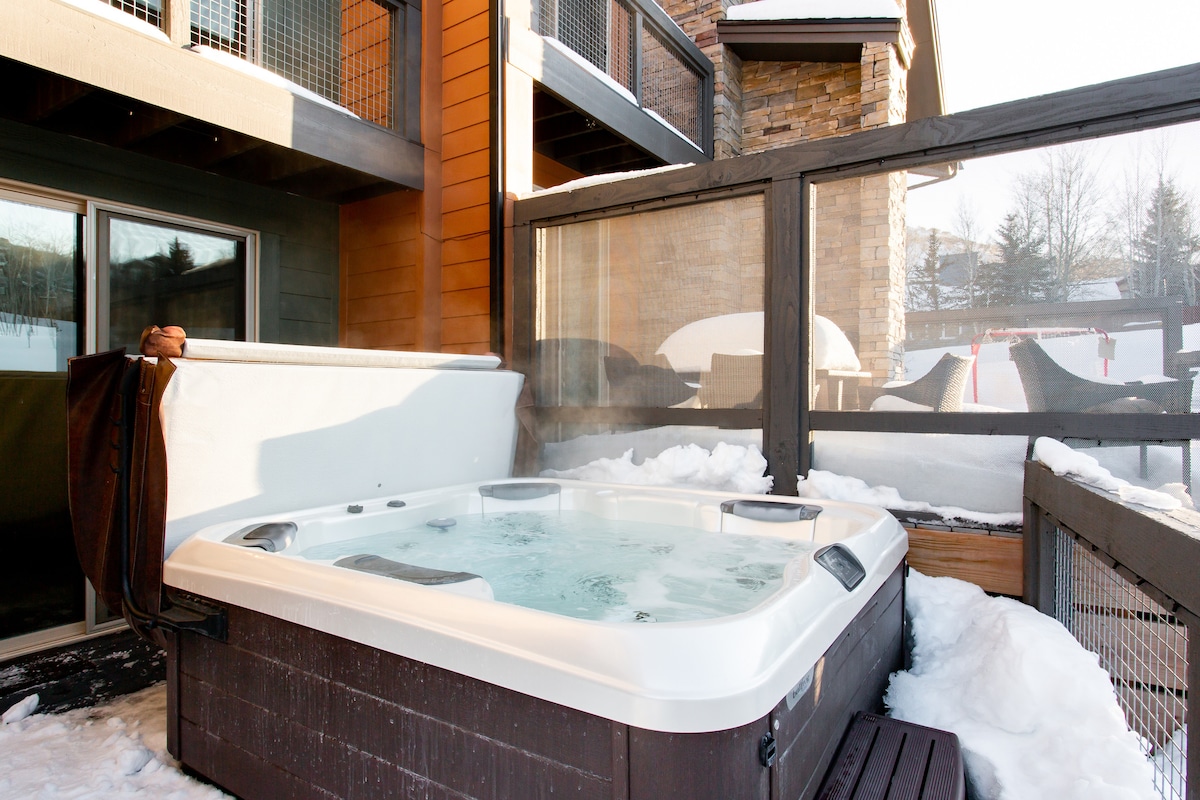 A private hot tub is situated on a deck surrounded by snow, with steam rising gently from the warm water. The area is enclosed for privacy, featuring wooden fencing and a glimpse of outdoor seating in the background.