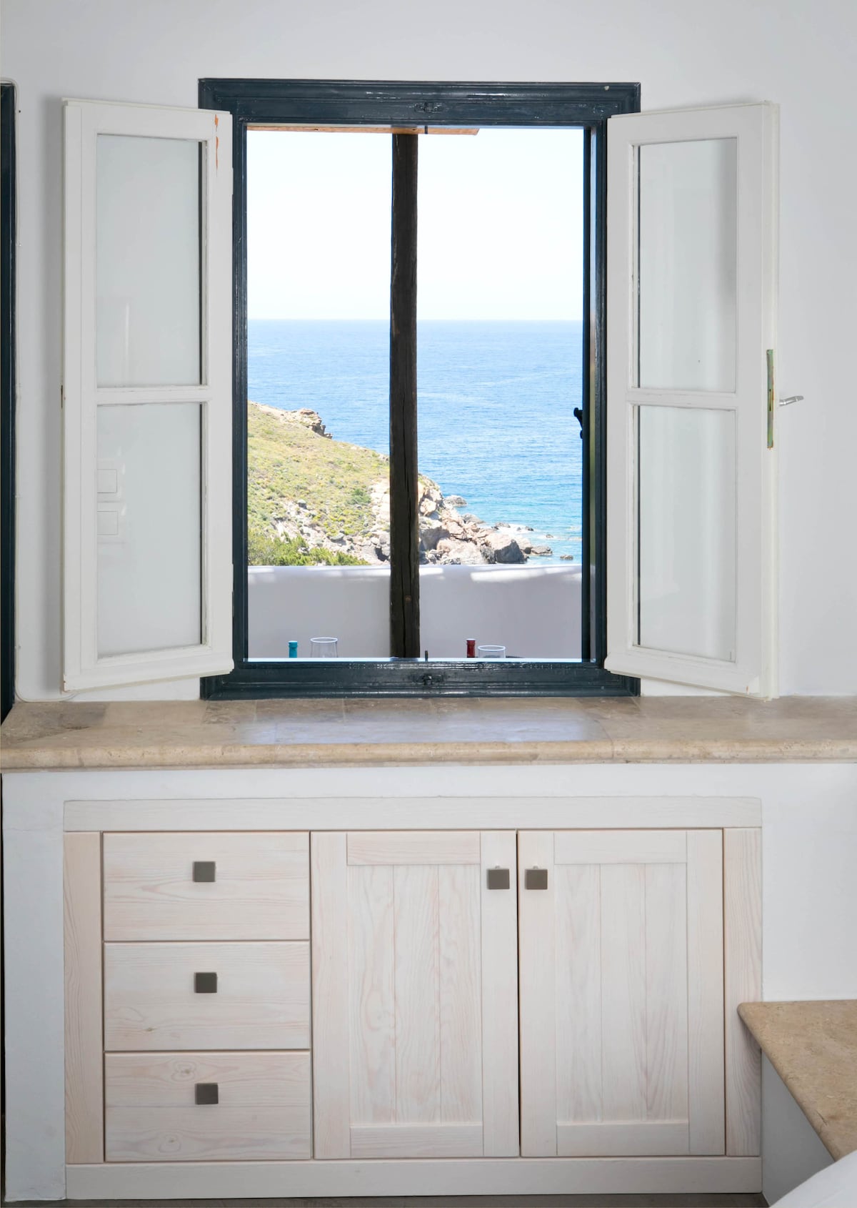 A window with dual opening shutters offers a clear view of the serene sea and rocky coastline. Light-colored cabinetry is positioned below, enhancing the space's airy feel. The coastal scenery can be seen in the distance through the open window.