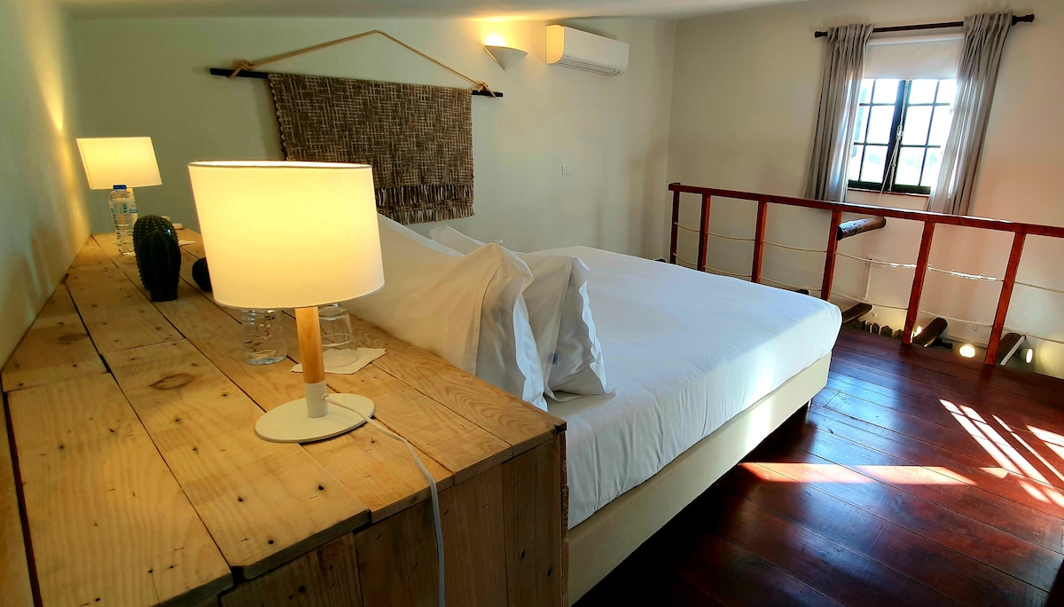 The bedroom features a spacious bed with crisp white linens, accompanied by a wooden bedside table holding a lamp. Natural light streams in through a window framed by light curtains, illuminating the warm wooden flooring and creating a serene ambiance.