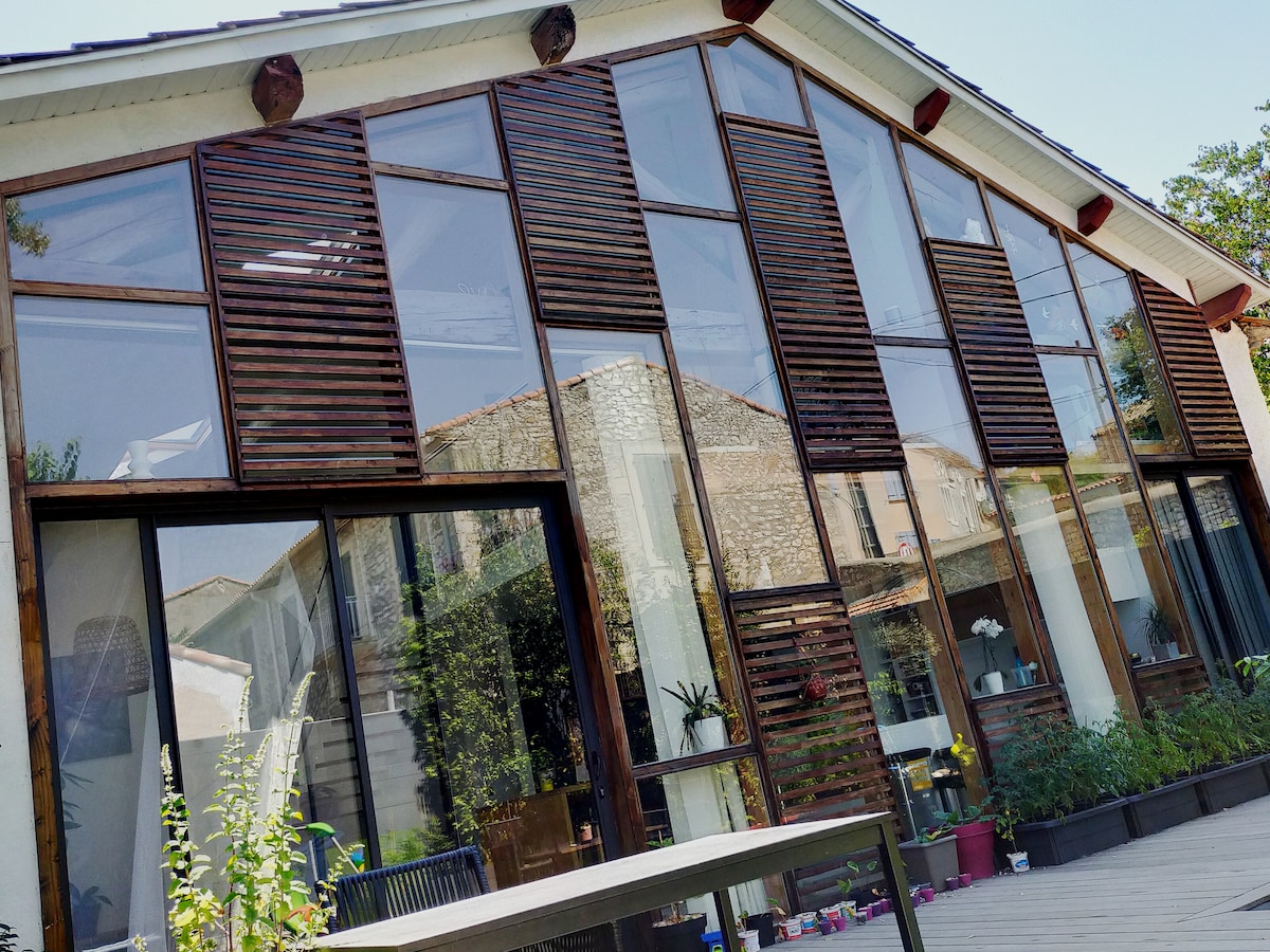 A modern house facade features large glass windows framed by wooden shutters. The design allows for ample natural light while providing privacy. Visible plants and outdoor furniture create an inviting transition from indoors to the surrounding garden.
