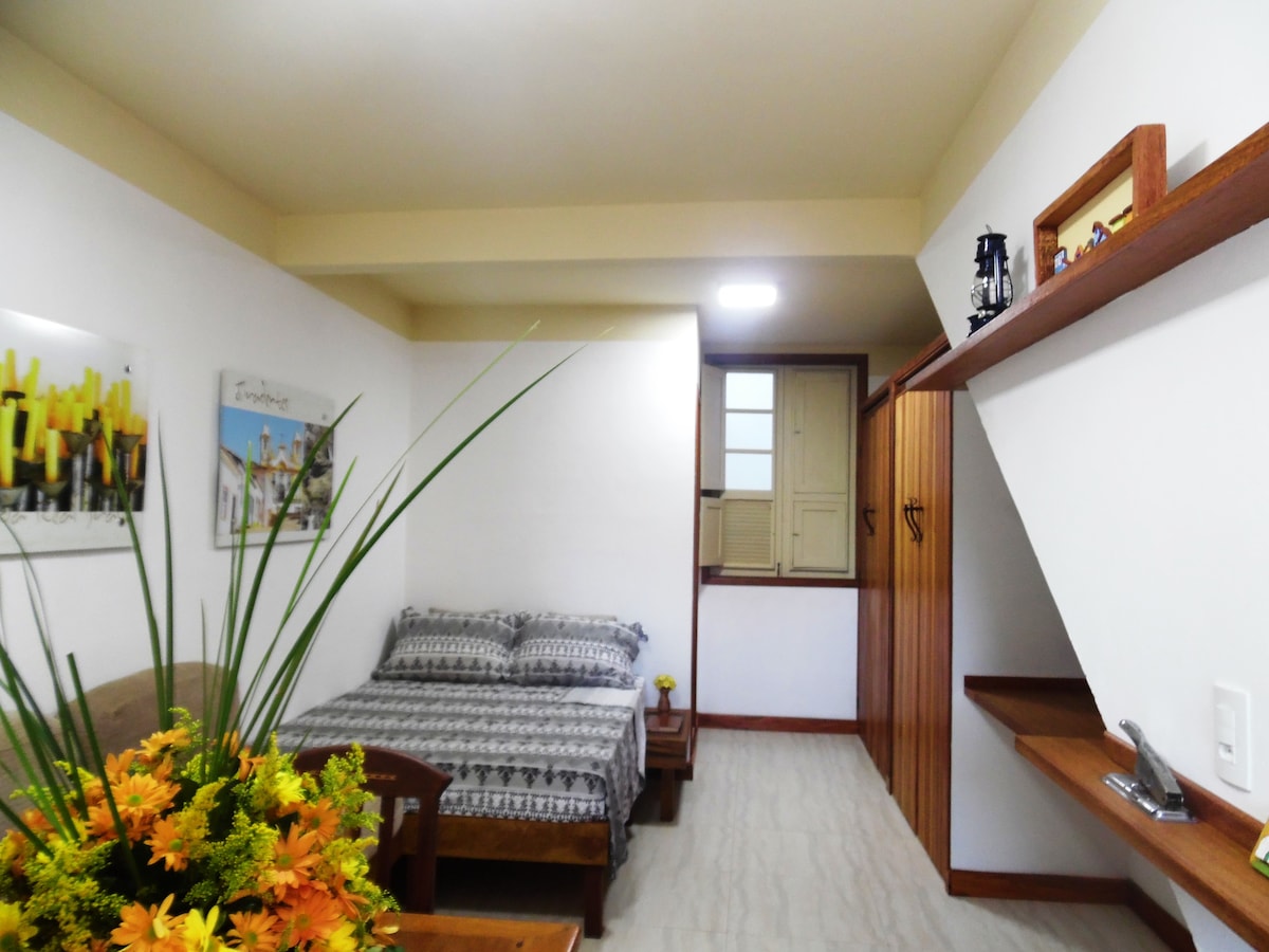 A spacious bedroom features a comfortable bed with a patterned blanket. A vibrant arrangement of flowers adds a pop of color in the foreground. Wooden shelves and a wardrobe provide storage, while natural light streams through the window, illuminating the tidy space.